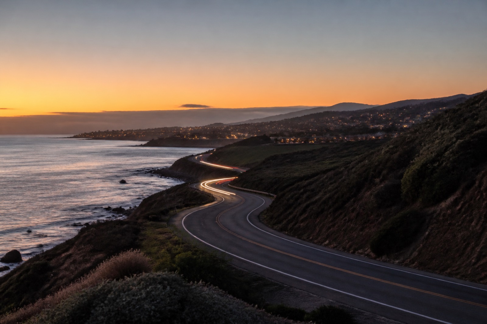 Coastal highway at dusk with suburban hills and city lights
